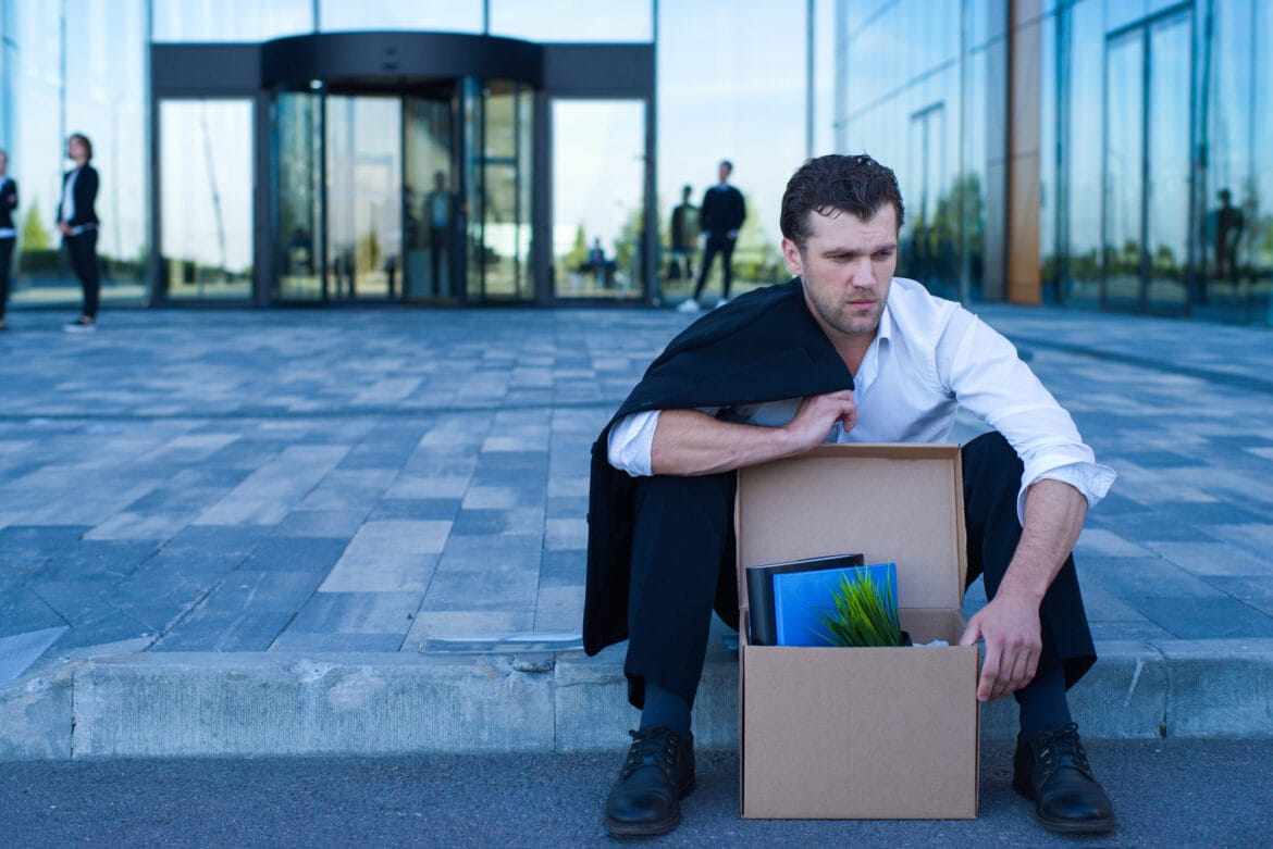 Fired business man sitting frustrated and upset on the street near office building with box of his belongings, he lost job